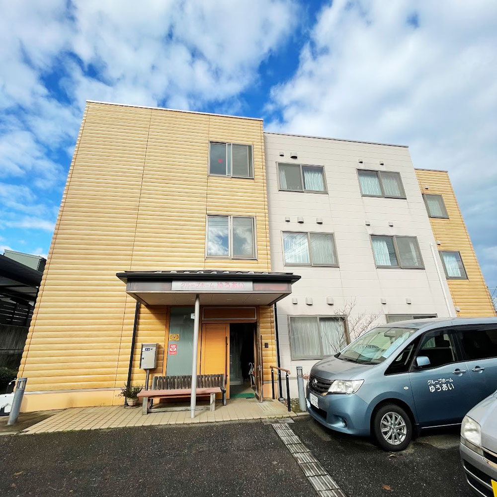 木製サイディングと白壁の介護施設の建物外観。駐車場と玄関周りが見える。